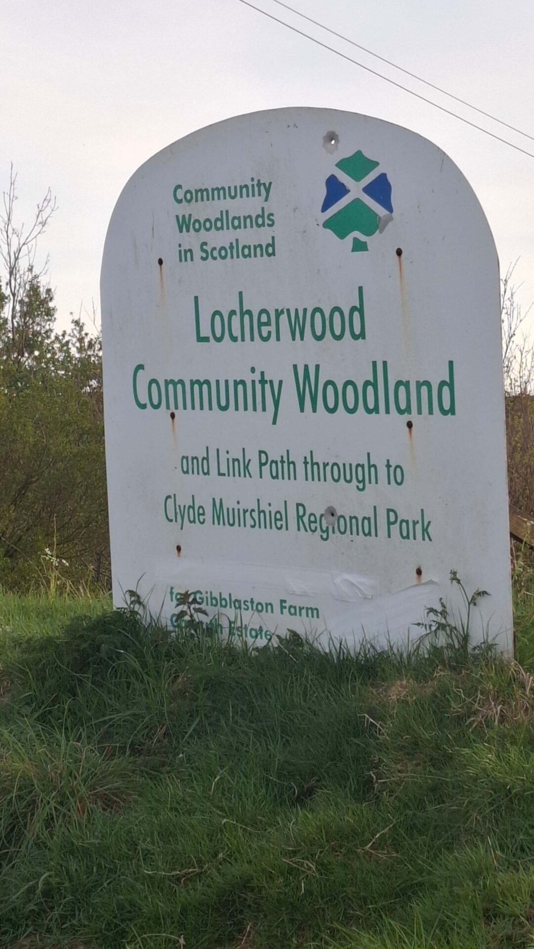 A white outdoor sign on a grassy verge, providing information about Locherwood Community Woodland and its connection to Clyde Muirshiel Regional Park.