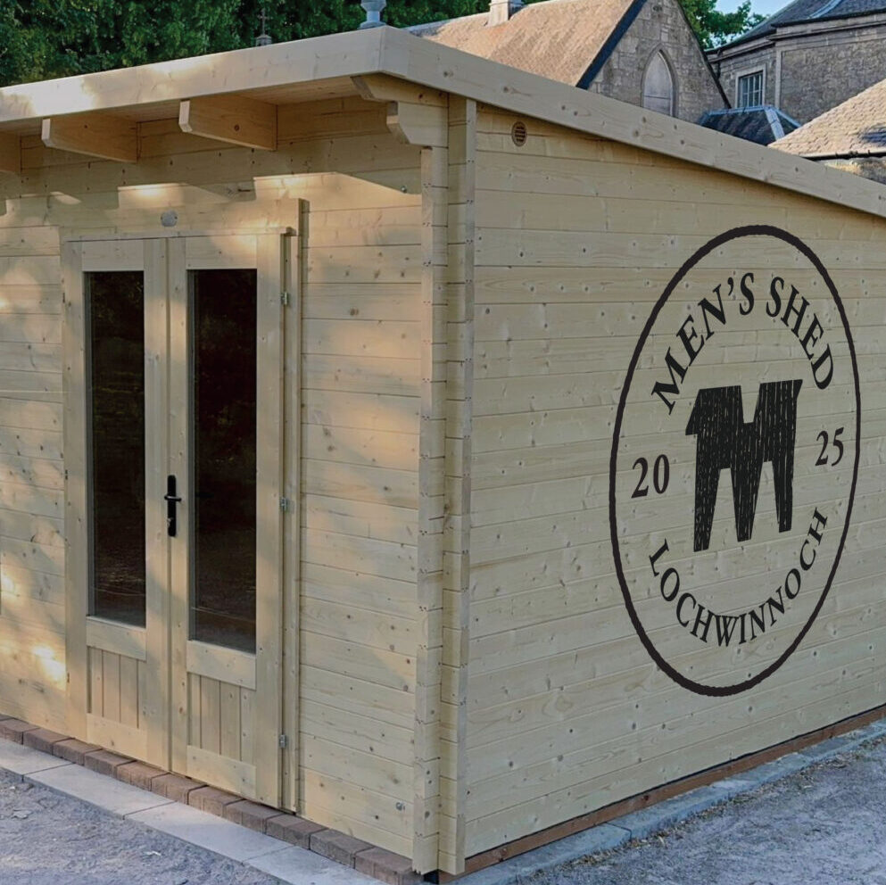 The men's shed, constructed from timber with a sloped roof, 3 narrow windows and a double door, situated on a gravel surface surrounded by trees in Lochead Gardens