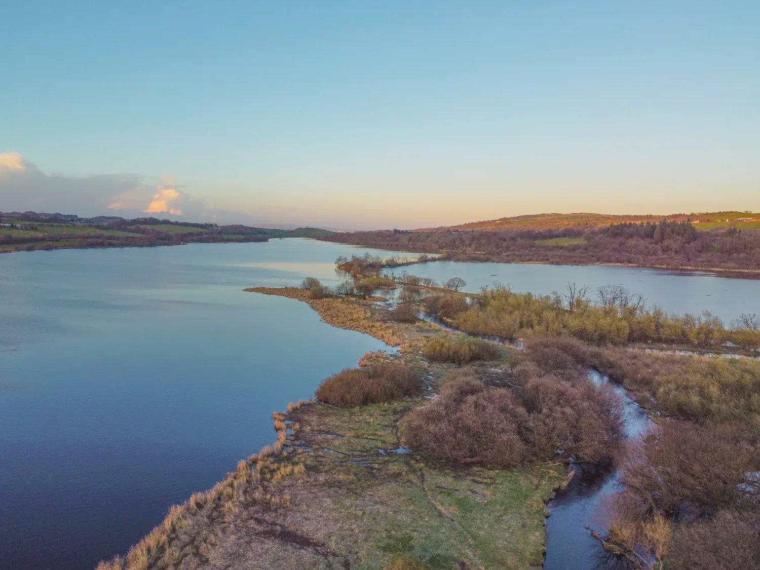 Aerial view of Castle Semple Loch at sunset, showing calm blue water, marshy wetlands with low bushes, and distant rolling green hills under a soft blue and orange sky.