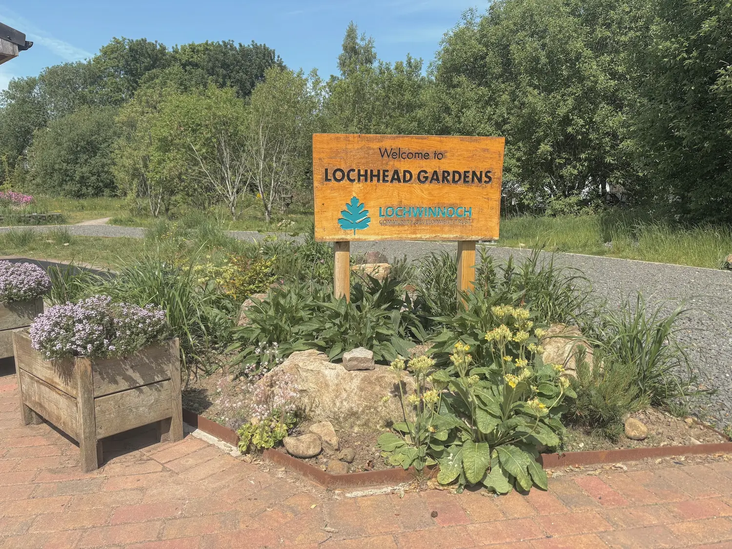A rustic wooden entrance sign reading "Welcome to LOCHHEAD GARDENS" with the Lochwinnoch Community Development Trust oak tree logo, set within a landscaped rockery garden with paths and trees behind it.