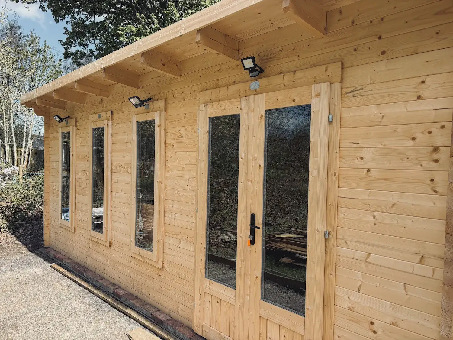 A brand-new, light-coloured wooden cabin workshop with double glass doors, three narrow vertical windows, and sloped roof.