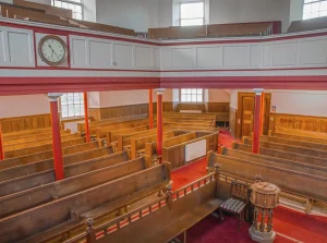 Interior view of a church sanctuary, showing rows of empty wooden pews on a red carpeted floor, red support pillars, and an upper gallery level with white paneling and a wall clock.