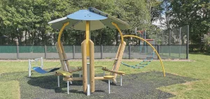 An outdoor modern park shelter for teenagers, featuring a blue circular canopy roof, timber and curved steel pillars with integrated seating, and attached hammock-style swings on a rubber mat floor.